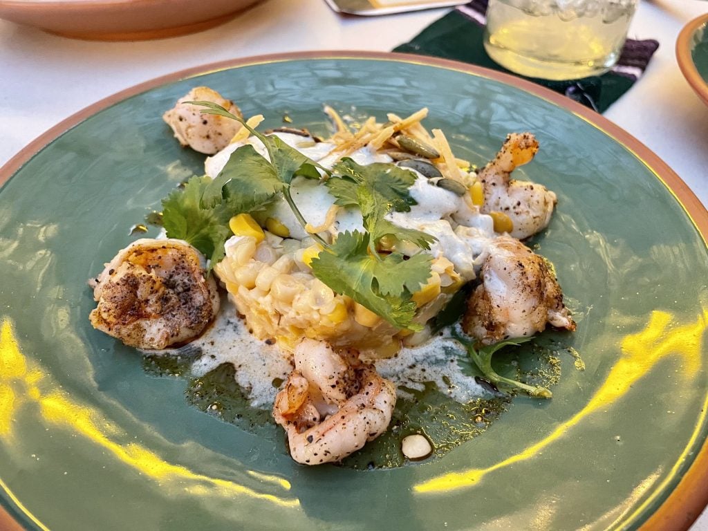 A plate with grilled shrimp resting around a pile of corn topped with foam and seeds.
