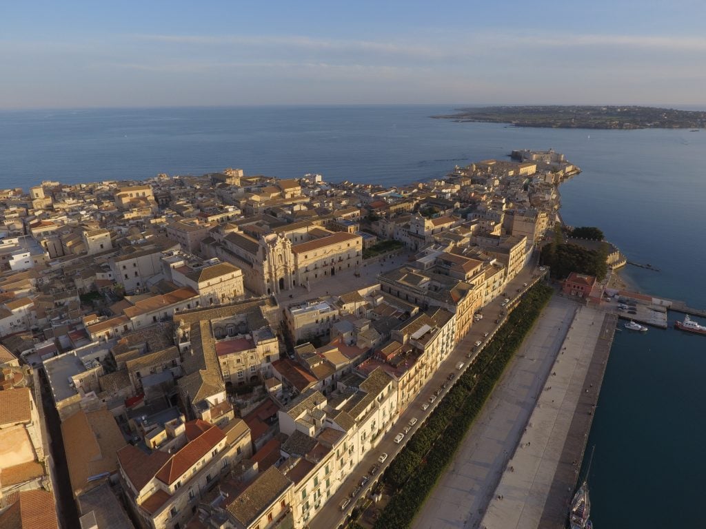 An aerial view of Ortigia Island in Siracusa, covered with rows of sand-colored stone buildings, one pointy end finishing in the bright blue sea.