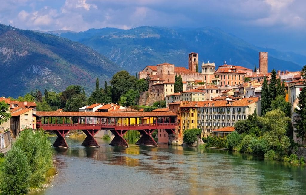 A beautiful small Italian town perched on a river, an old-fashioned wooden covered bridge crossing it, mountains in the background.