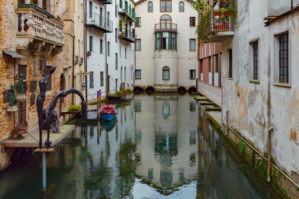 An Italian city of white buildings with a still green canal in between them, looking like Venice.